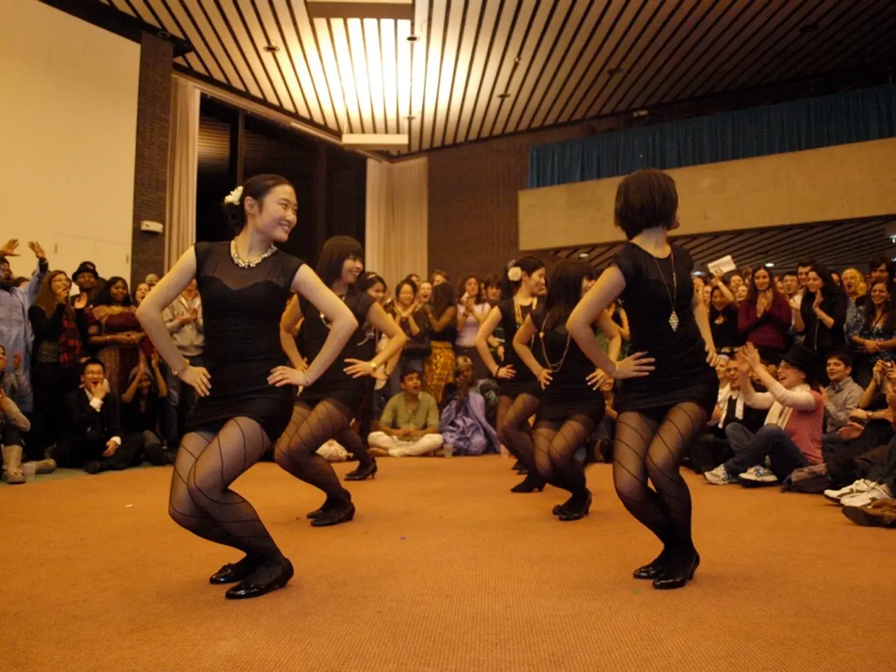In this image we can see five girls are dancing, they are wearing black color dress. Behind so many...